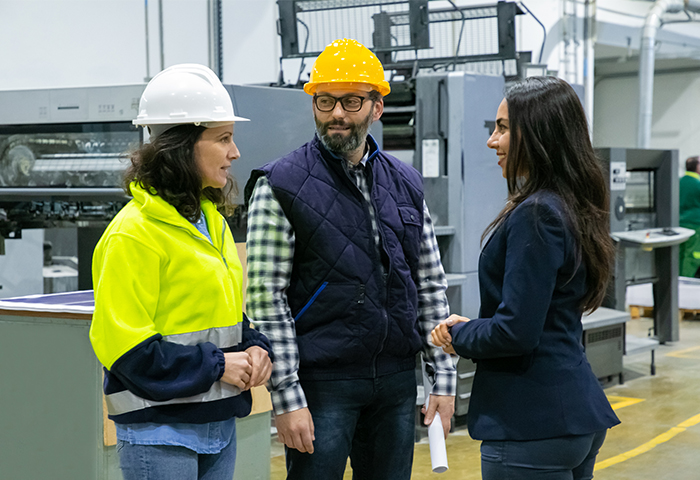 A hypothetical Alta Impact employee talks with two people wearing hardhats while standing in an industrial facility.